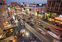 Downward view on a well-trafficked street surrounded by mid-rise buildings. On one side of the street are ceremonial stars embedded in a polished sidewalk.