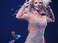 Britney Spears wearing a nude dress with crystals and is holding her arms at the height of her own head.