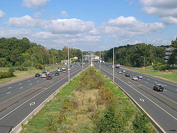 Both sides of a highway with a grass plot in the middle of the roads. Street lamps surround the middle and several cars are on the roads. The roads have an HOV diamond on them.