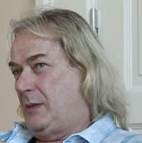 The head of a white man with long, blonde hair wearing a blue, button-down shirt in front of a white door.
