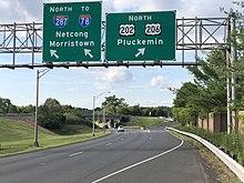 A multilane highway in a suburban area at a split, with two green signs over the road. The sign on the left reads north Interstate 287 to Interstate 78 Netcong Morristown with an arrow pointing to the upper right and the sign on the right reading north U.S. Route 202/U.S. Route 206 Pluckemin.