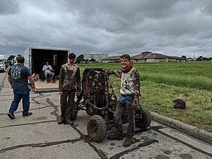 Baja SAE vehicle and two drivers, all completely covered in mud. Ominous rainclouds loom in the background, as well as vehicle trailers and a field of waving grass.