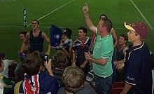 Group of fans chanting for their team in stadium stands adorned in Australian flags and various football paraphernalia.