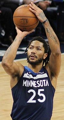 Hip length view of man raising the basketball to shoot free throw, with black moustache, beard, and short dreadlocks, thin white headband, wearing navy blue Timberwolves uniform