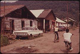 Three children bicycling on a dirt road in front of "substandard housing"