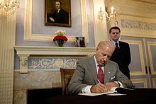 Joe Biden sits at a desk, writing in a book