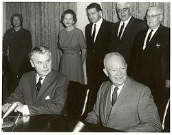 Diefenbaker and a smiling bald man in a suit sit at a table. Two women and two men stand behind them.