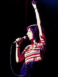 A female singer, Patti Smith, singing into a microphone at onstage performance. She holds the microphone with one hand; the other is raised up and in a closed hand.