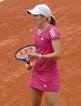 A blond-haired women is wearing a neon-pink shirt and white skirt, and is reaching to hit a one handed backhand