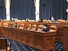 Several individuals in professional atire sit in leather chairs behind two rows of ornate wooden stands.