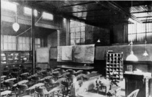 A black and white image of a large room with may chairs in front of a desk and charts. There are bars on the windows.