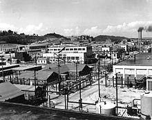 An array of industrial buildings with lots of power poles and wires, with a pair of smokestacks in the background