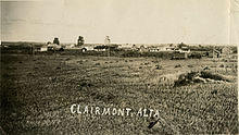 A black and white image of a rural town with three elevators and town buildings from an adjacent hay field