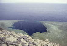 View of the coastal waters from the top of a hill, showing an approximately circular hole in the shallow coastal reef tangent to the deeper water offshore.