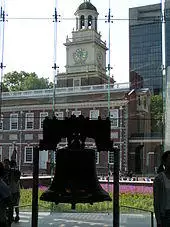 The Liberty Bell hangs in a glass-backed structure, with a brick, 18th-century building with a steeple visible in the background.