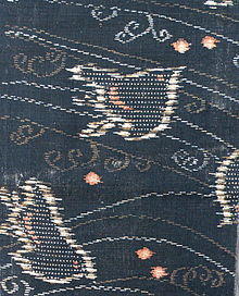 A close-up of a piece of kasuri-dyed fabric with a woven design of plovers and stylised waves on a dark indigo blue-green background. The waves are woven in both white and brown, and the plovers are woven in a mixture of white, brown and pink.