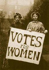 Two woman carry a sign reading "Votes for Women".