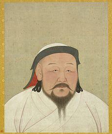 A head-shot style portrait of a middle aged man with flush cheeks and a black beard that extends from ear to ear, and is part in the middle, as well as a long mustache extending out horizontally rather than down. He is wearing a white robe and a black trimmed white had with a red inside lining.