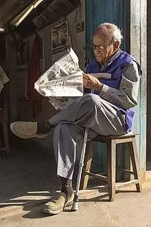 Photograph of a bespectacled man sitting on a stool with his legs crossed reading a newspaper in the morning