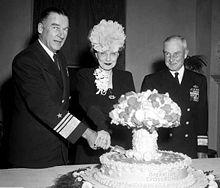 A man in naval uniform and a woman wearing a hat cut into a cake labeled Operation Crossroads, and shaped like a mushroom cloud, while another naval officer looks on.