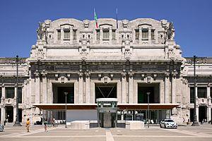 An ornate and extensively detailed gray stone classical portico with four sets of twinned columns at the top of which the Italian and European Union flags hang limply; above is clear blue sky. In front is a more modern one-story pavilion with an elevator in front of it inside a circular wall. Some people can be seen walking around.