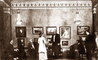 Scene from a play, with young woman standing in a smart drawing room addressing several seated men