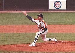 A man in a white baseball jersey with "ASTROS" on the chest and orange and yellow stripes on his shoulders pitches a baseball with his right hand.