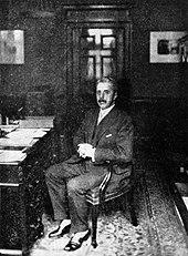 man in late middle age, full head of greying hair, moustached, sitting at a desk