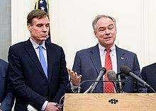 Two older white men in suits stand behind a podium while one talks.