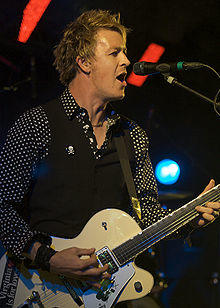 Cheney is singing at a microphone while playing his guitar. He is shown in right profile, he wears a dark shirt with white polka dots, covered by a sleeveless vest. Some stage lights are visible behind him.