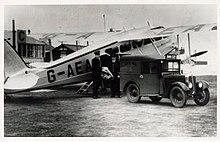 Photograph of a truck backed up to the side of an aeroplane with two men holding a mail bag