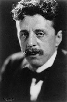 Middle-aged white man with full of wavy dark hair swept into a substantial quiff. He has a neat, medium-sized moustache.