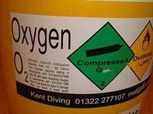 The white adhesive plastic label displays the gas name, Oxygen, and the chemical symbol O2 with a block of small text on the left side describing the hazards of the contents, then a green diamond symbol for compressed gas and a yellow diamond for oxidising agent.
